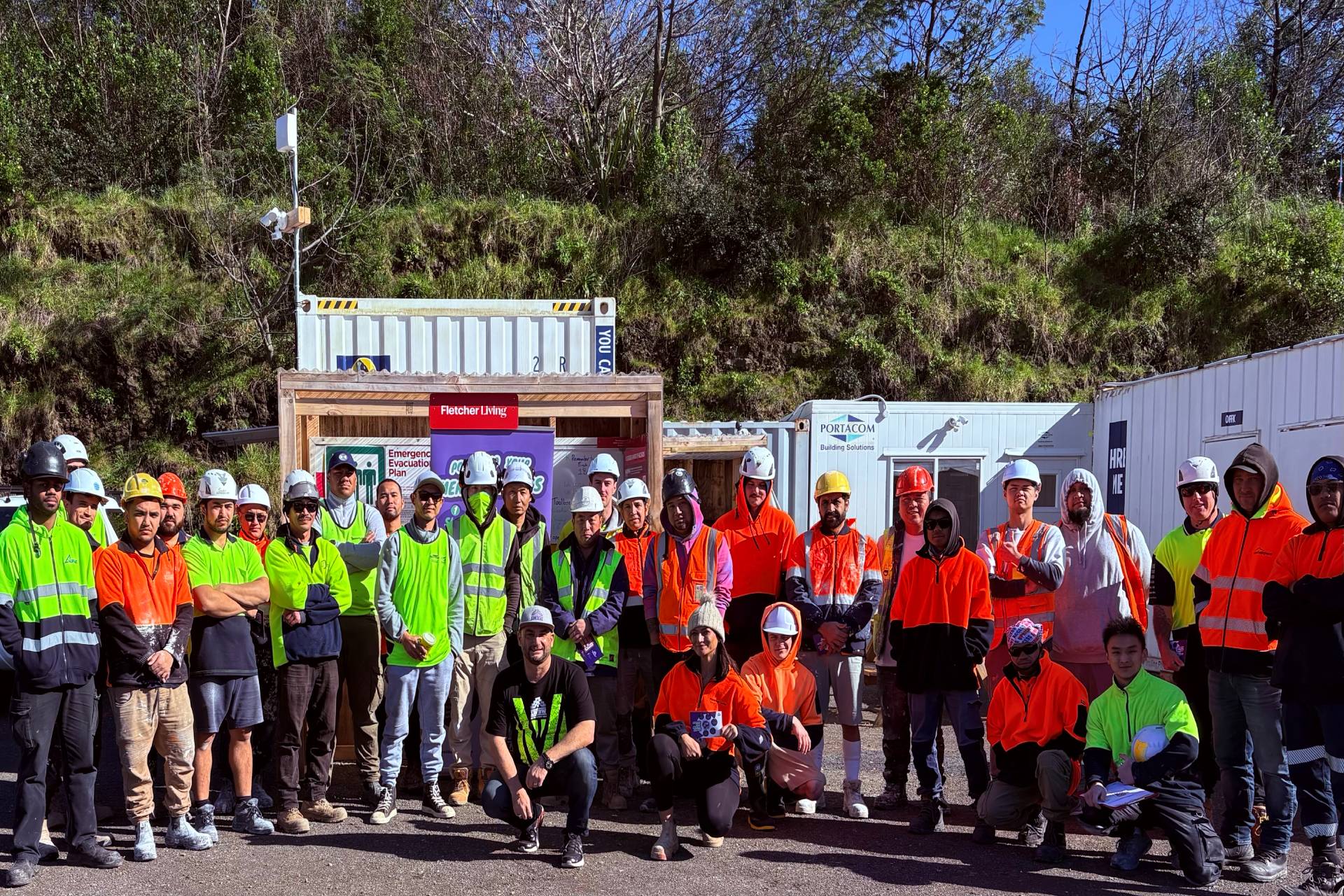 A large group of construction workers wearing safety vests and helmets pose together outdoors at a job site.