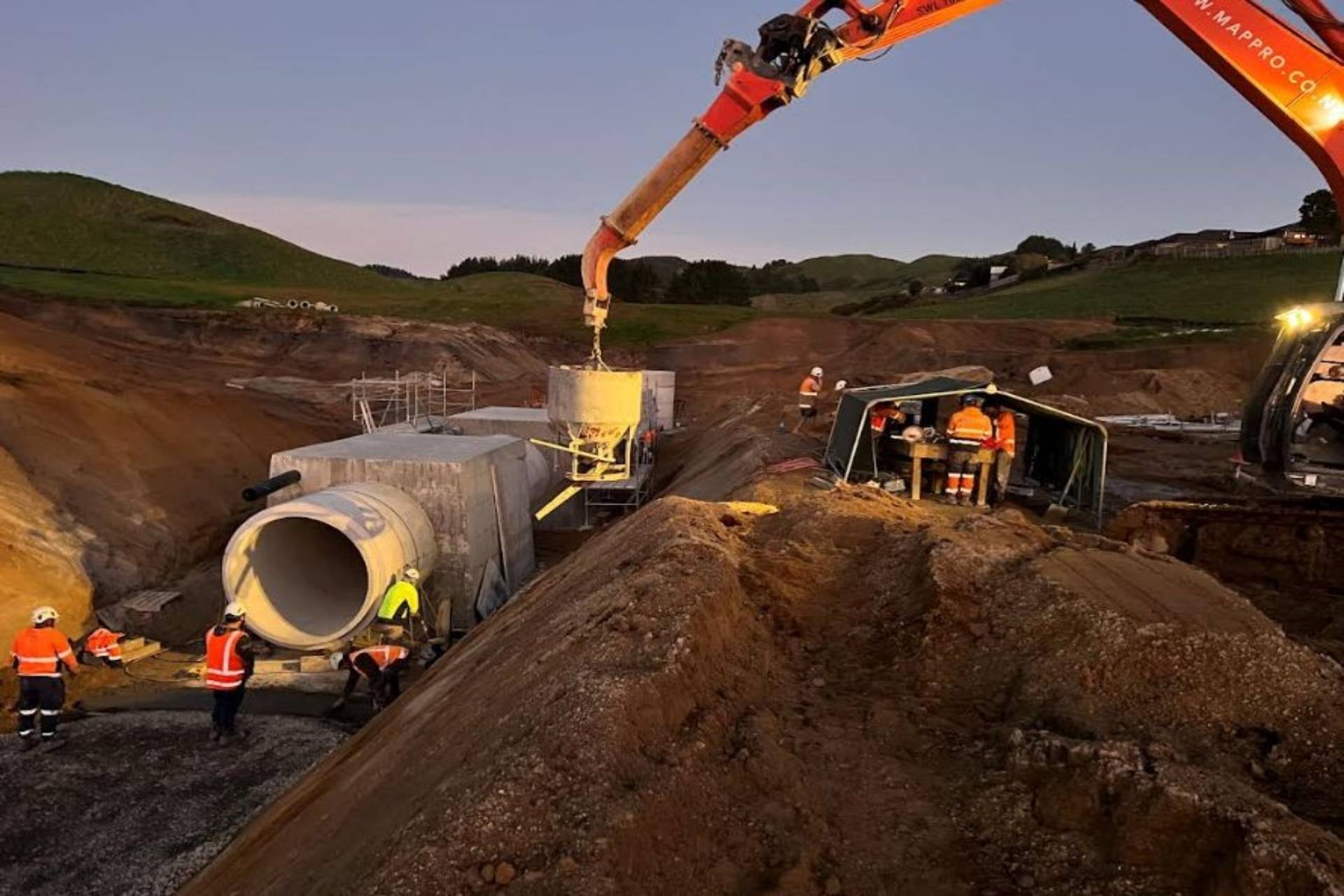 Workers pouring concrete into large drainage pipes at a construction site during dusk